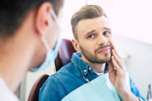 Man with worried look, holding hand to cheek in pain due to infected tooth, needing root canal.
